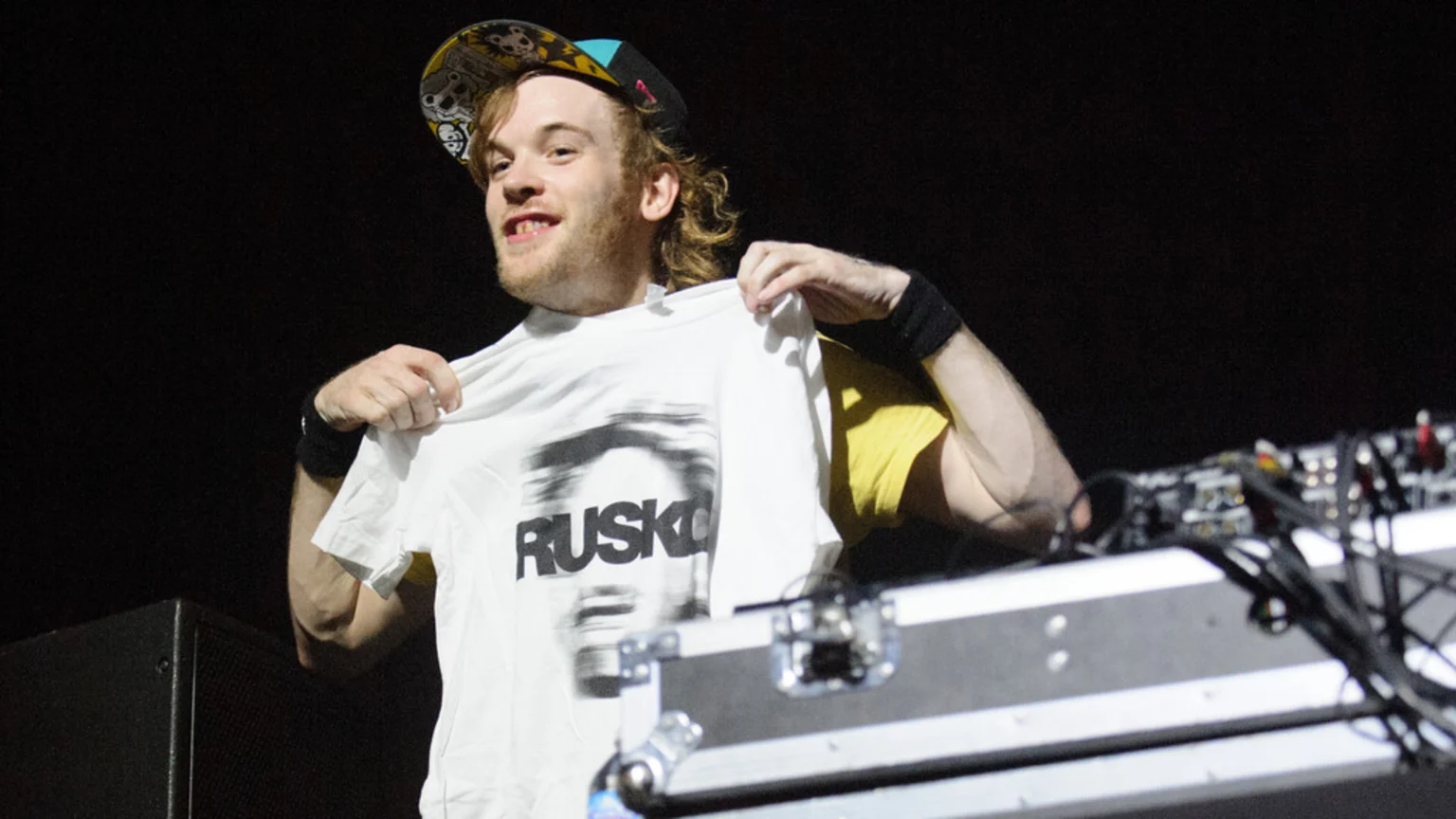 A smiling man, identified as Rusko by his t-shirt, holds up a white shirt with a blurry face graphic and "RUSKO" printed on it, standing in front of dark background.