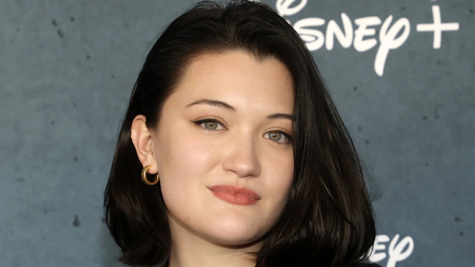 Close-up portrait of Isa Briones with dark hair, light eyes, and a slight smile, wearing a gold hoop earring, against a dark grey background with the Disney+ logo.