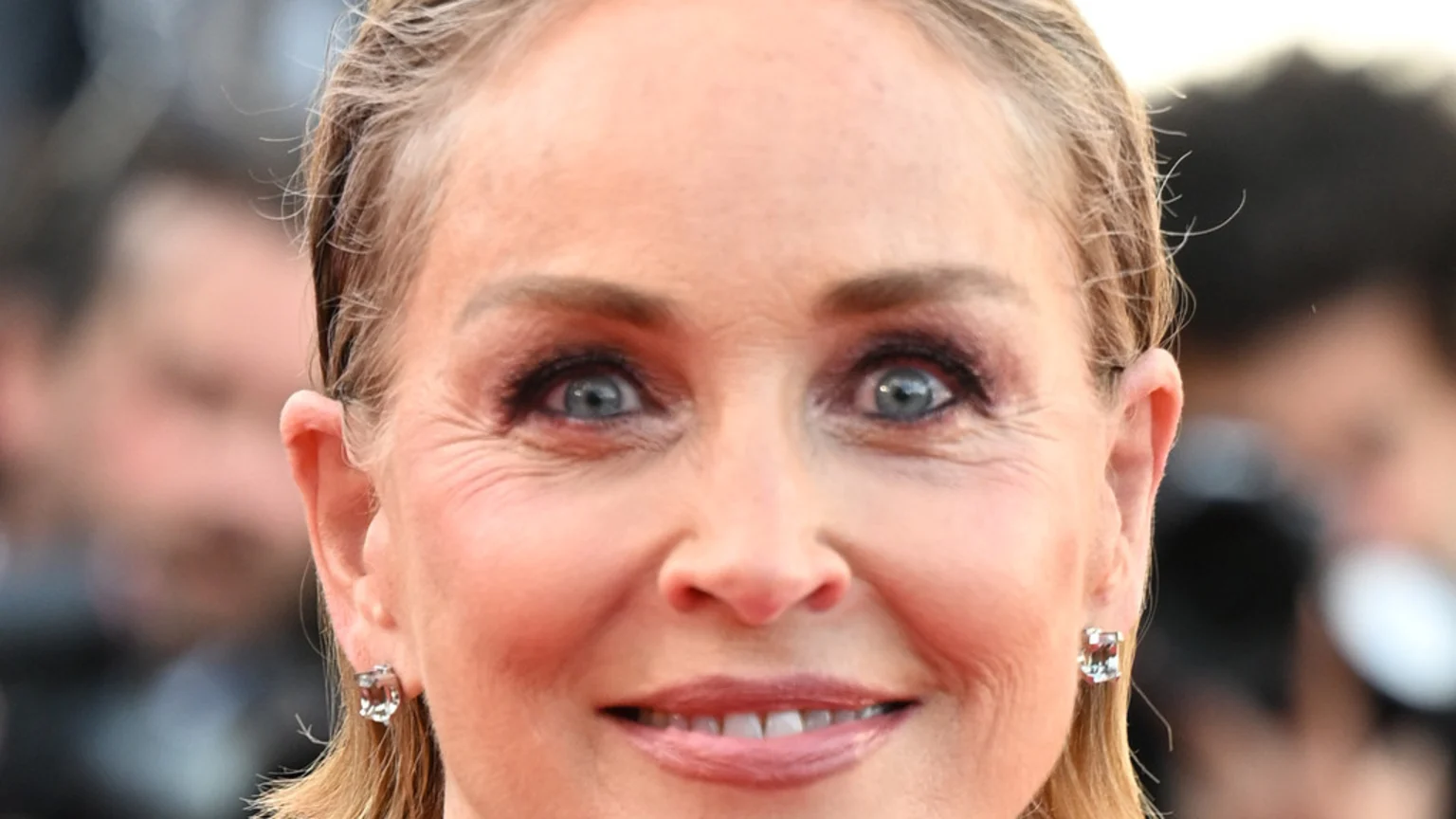 A close-up headshot of Sharon Stone smiling slightly and looking directly ahead, showing her blue eyes, light brown hair pulled back, and small stud earrings.