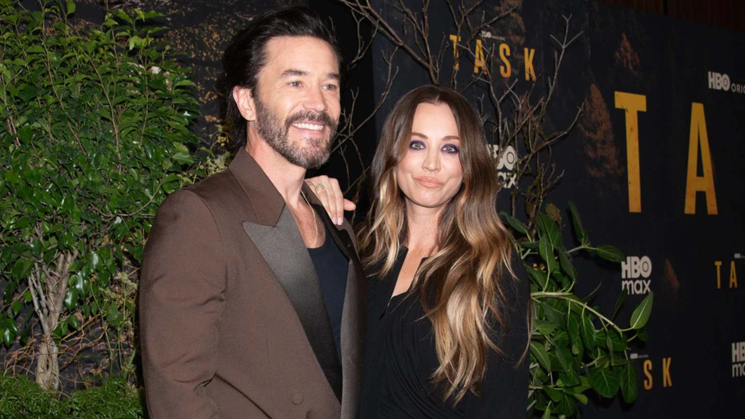 Two unknown individuals pose indoors at a TASK event premiere featuring HBO Original and HBO Max branding on a dark backdrop with green foliage and bare branches. Both wear formal attire and smile subtly.