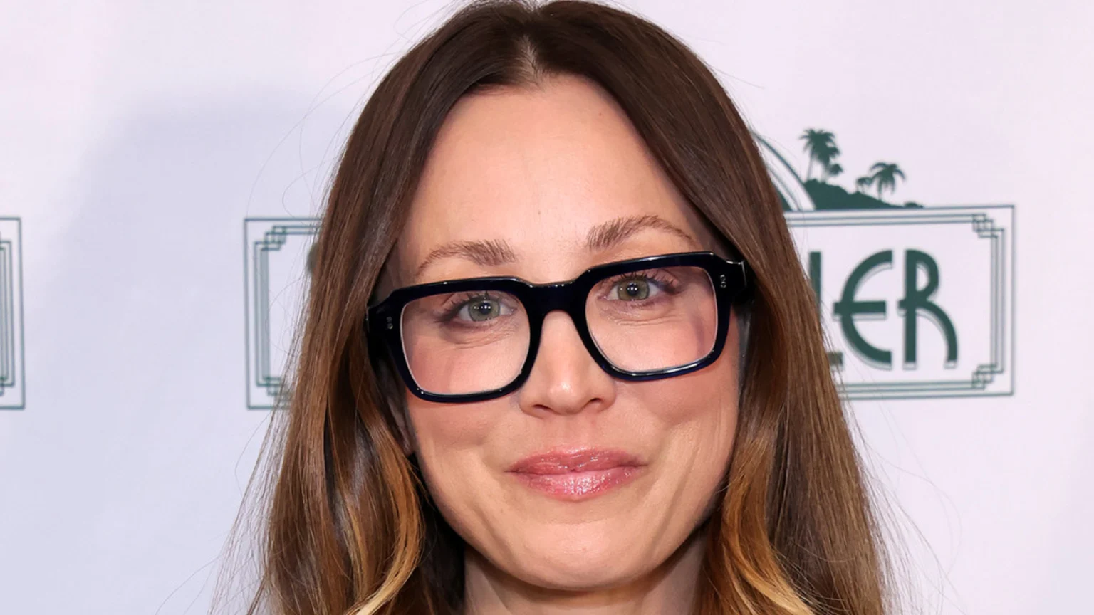 A close-up portrait of a middle-aged woman with long brown hair and dark, thick rectangular glasses, smiling gently against a white studio backdrop adorned with green graphic elements, including a stylized palm tree logo and geometric frames.