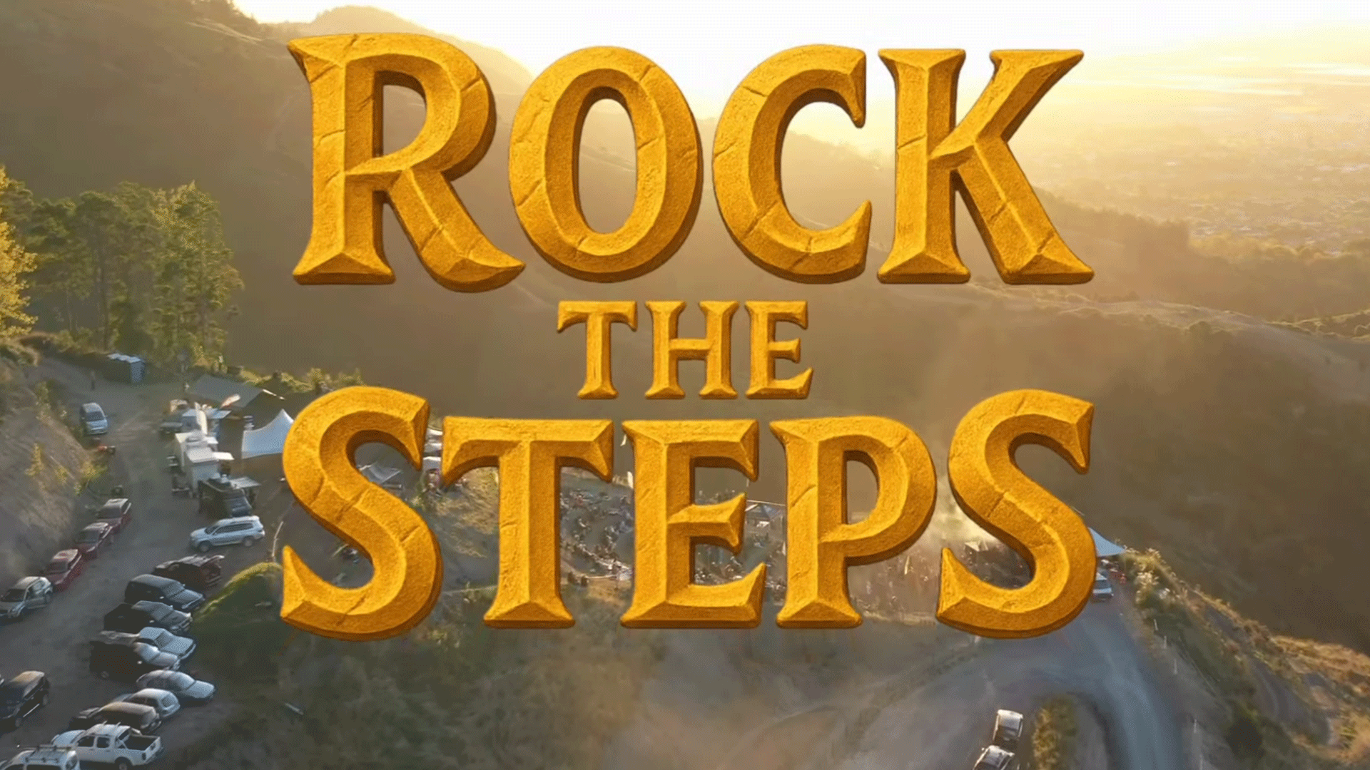 Golden "ROCK THE STEPS" text in a stone-like font overlaid on an aerial view of a hilly landscape with trees, winding roads, and parked vehicles under a warm, golden sunset sky.