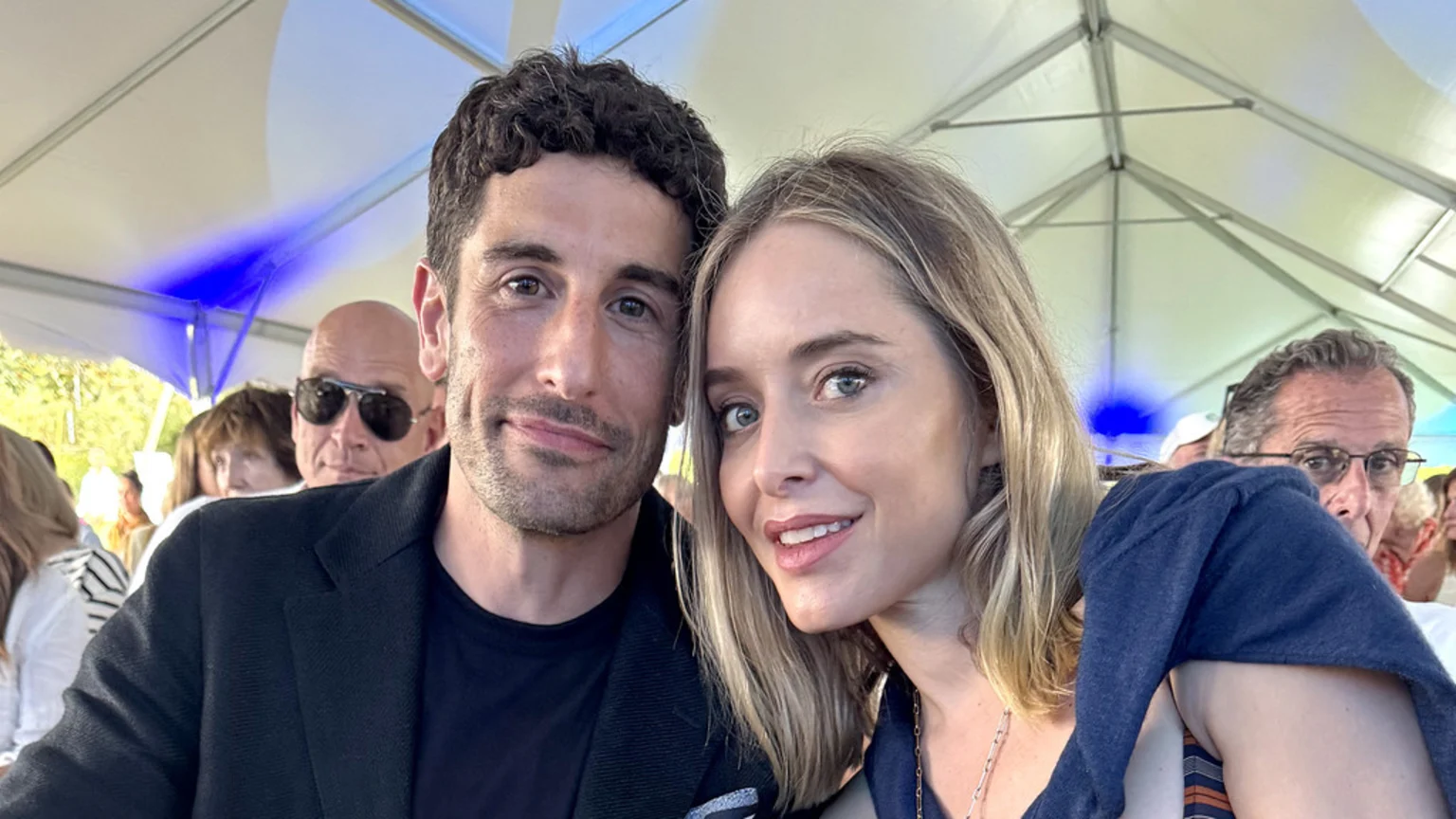 A man with dark curly hair and a woman with long blonde hair pose closely together, smiling at the camera, with other people and a white tent visible in the background.