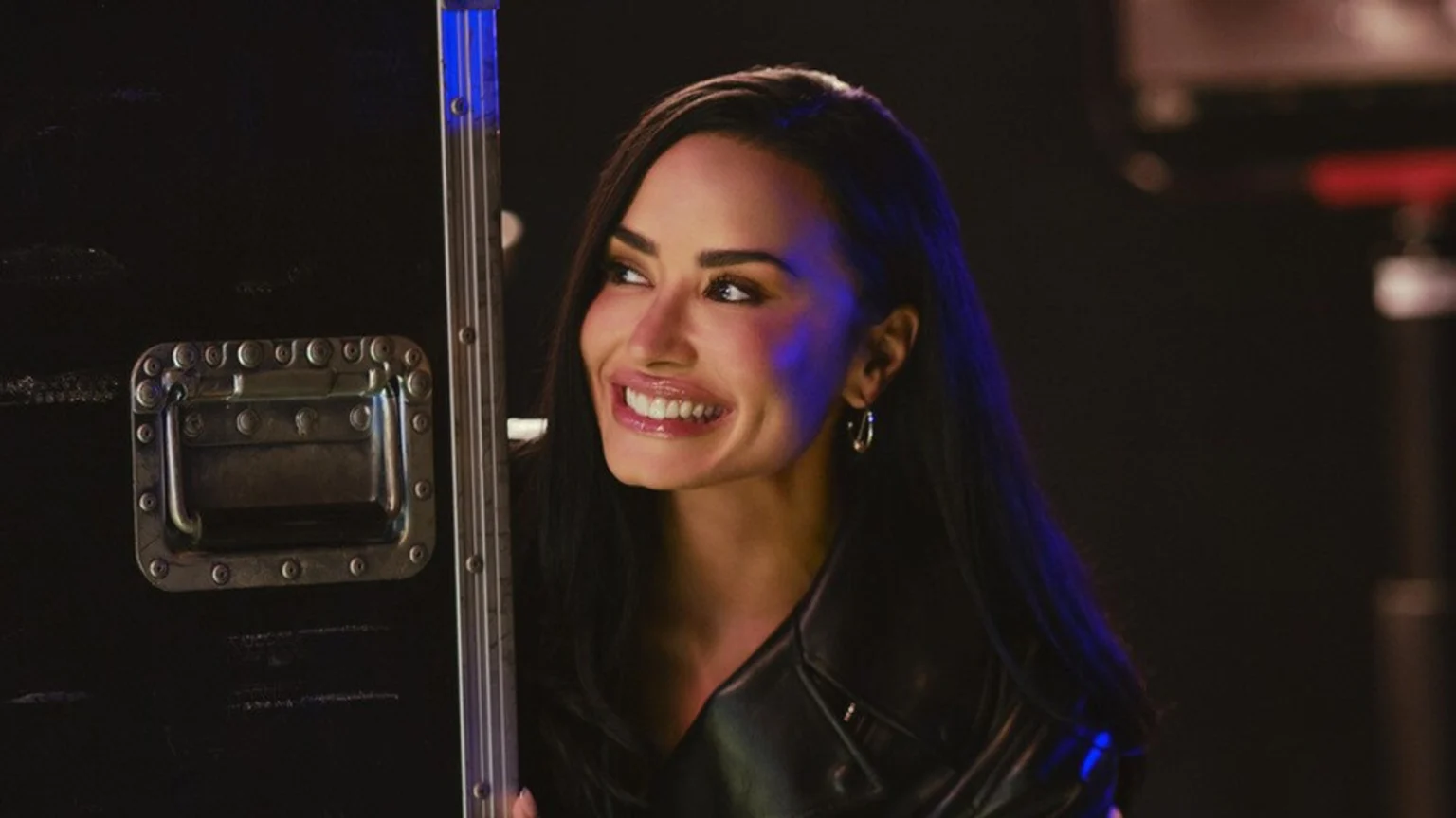 Demi Lovato with long dark hair smiles broadly, wearing a black leather jacket, looking leftwards as blue light reflects on her hair and a dark metal equipment case beside her.