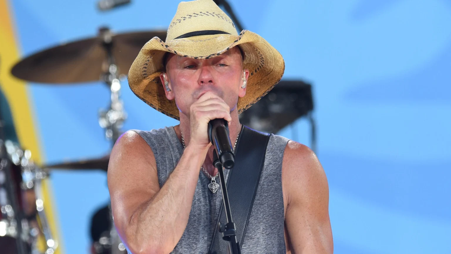 A man in a straw cowboy hat and grey sleeveless shirt sings into a microphone, with a blurry blue background showing musical equipment.