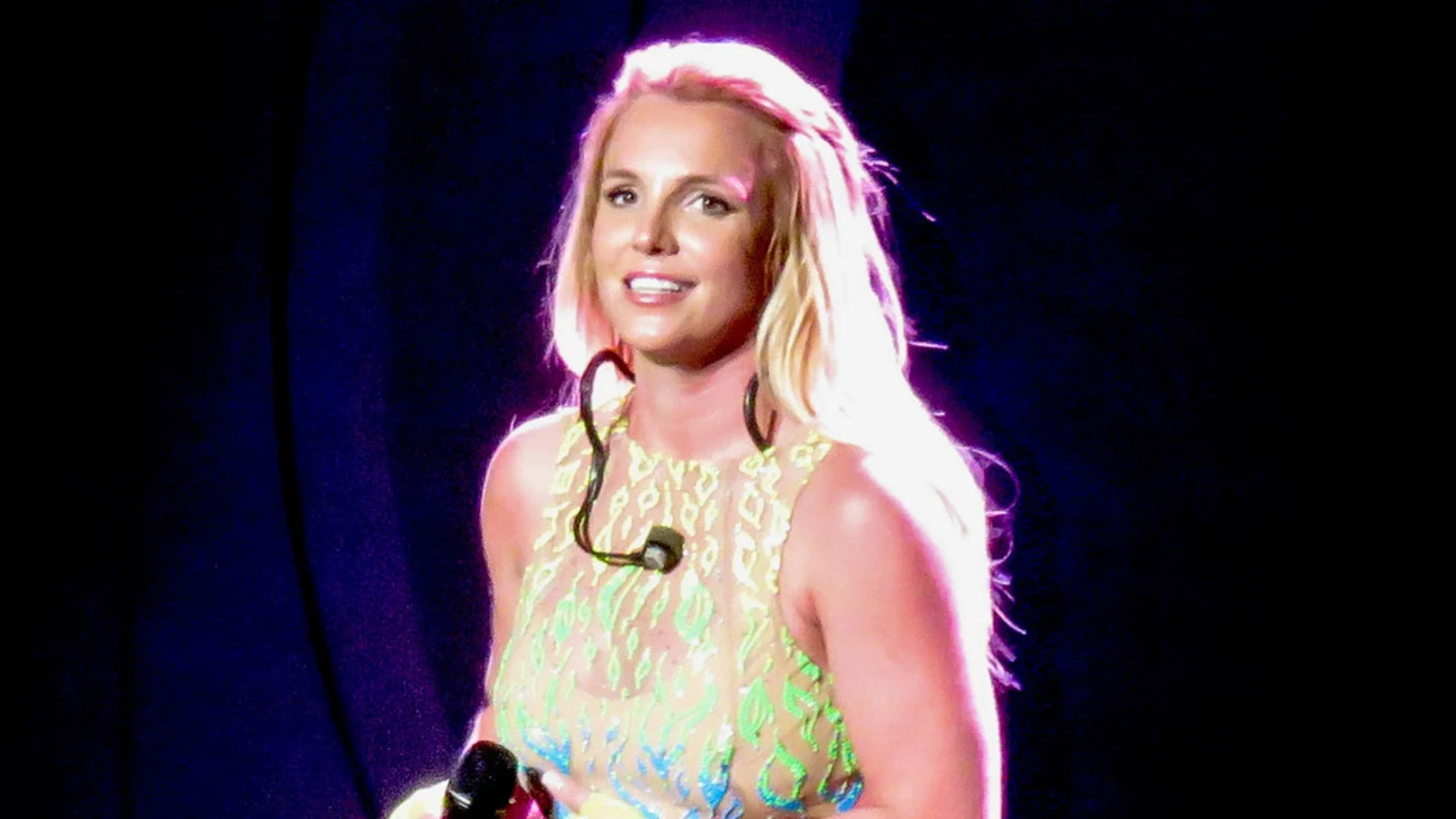 Britney Spears, with long blonde hair, smiles on stage while wearing a patterned sleeveless top and ear-mounted microphone, holding a handheld microphone.