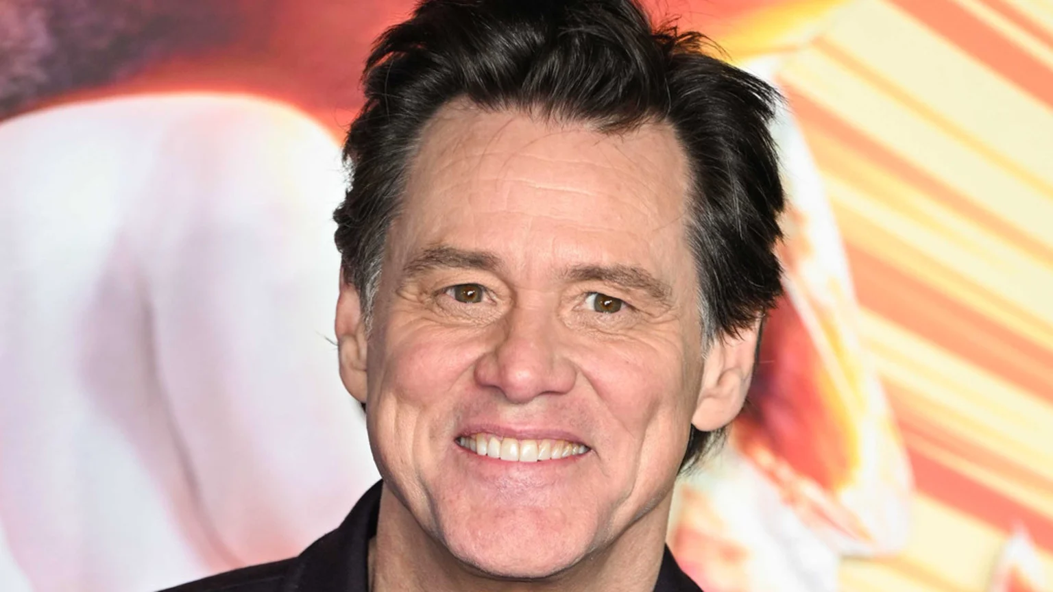 A smiling Jim Carrey looks forward, with dark hair and a blurred background of warm, abstract colors.