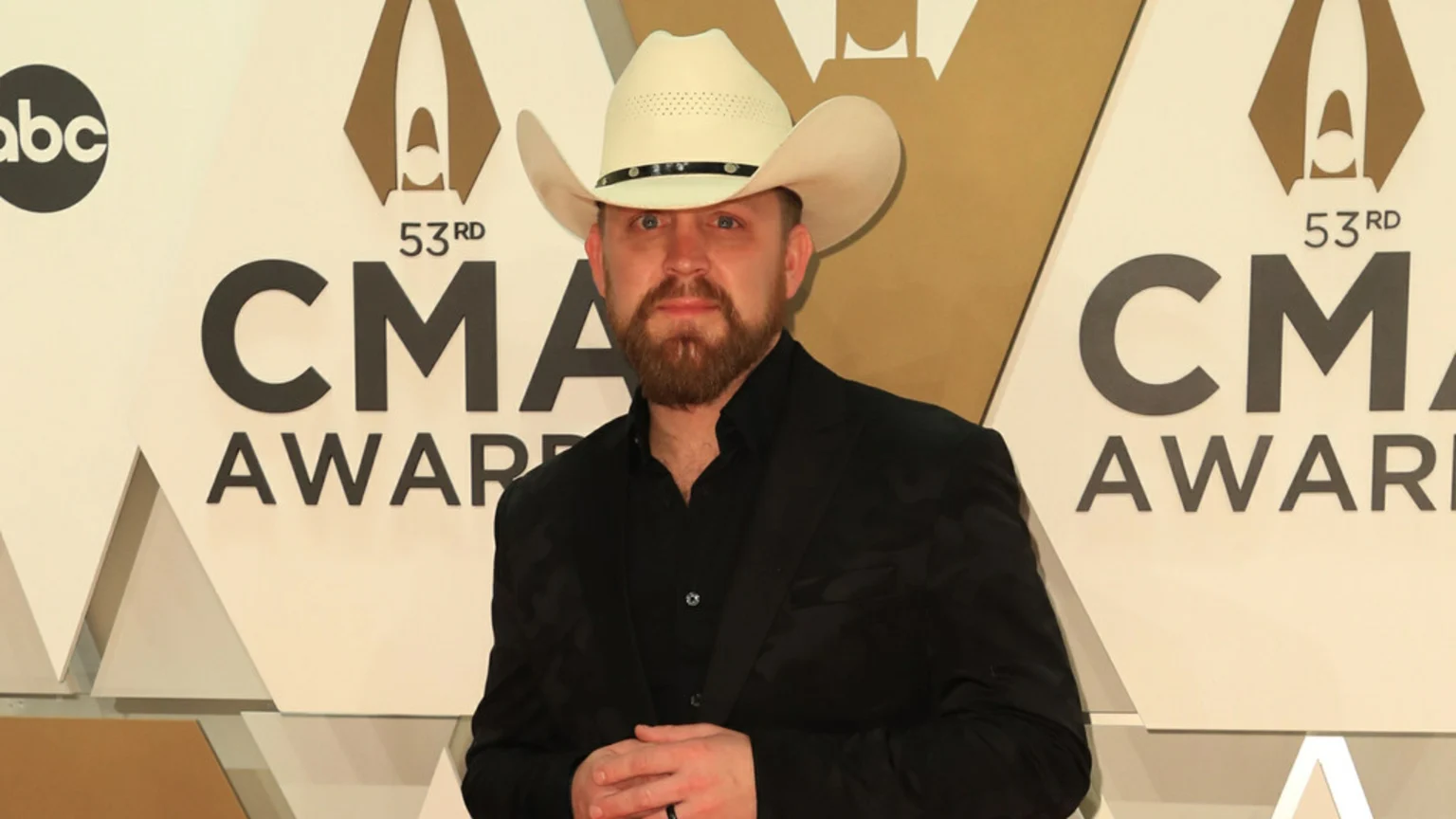 Country music artist Justin Moore, with a beard and white cowboy hat, poses in a black suit against a "53rd CMA Awards" backdrop.