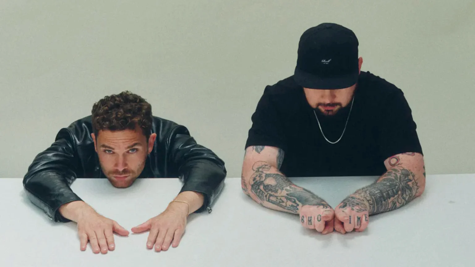 Two men sit at a white table; one in a leather jacket looks forward, the other in a black cap and t-shirt looks down, displaying tattooed arms and knuckles.