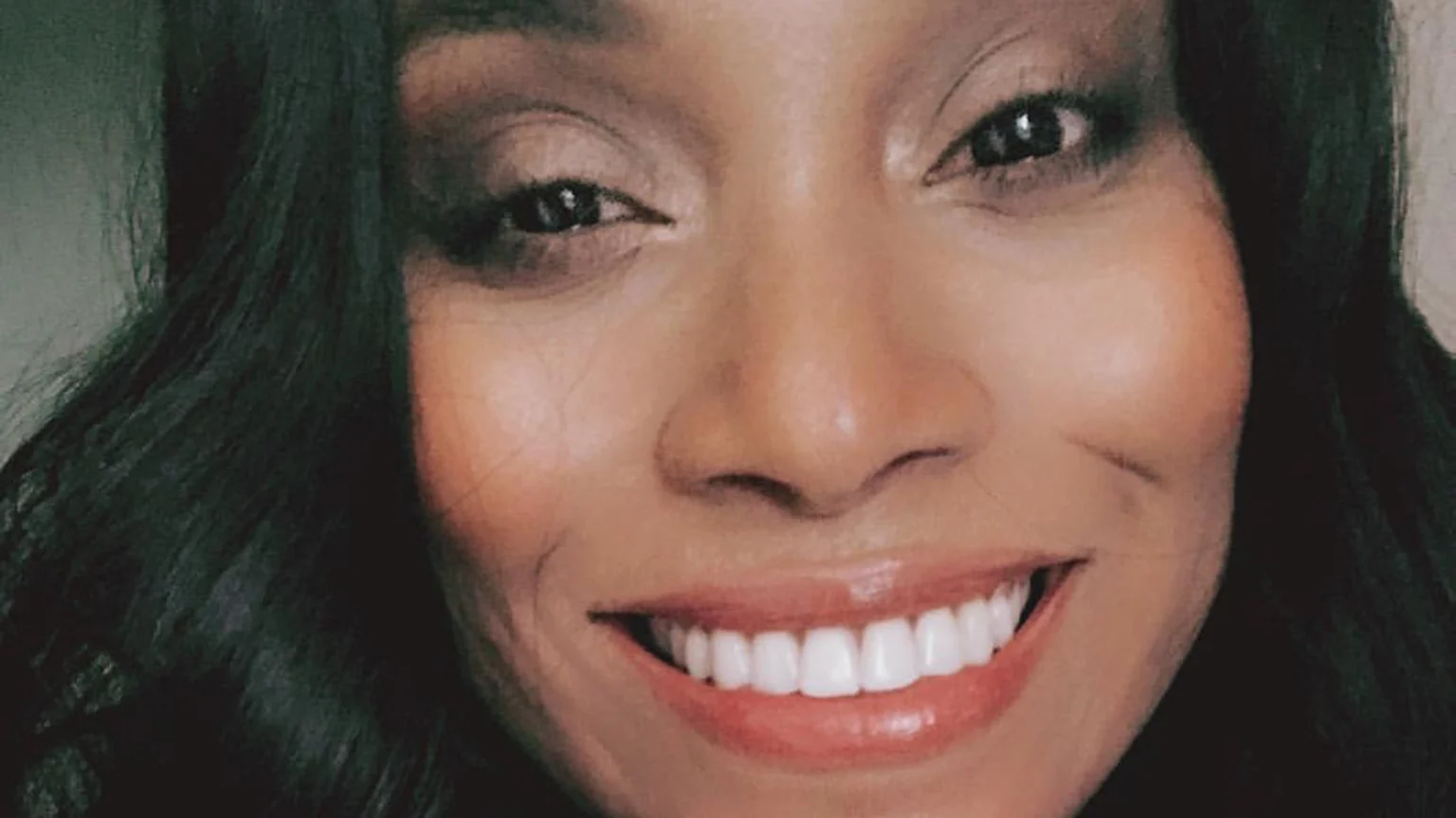 Close-up of a smiling woman with dark skin and long, dark, wavy hair, wearing makeup and looking towards the viewer.