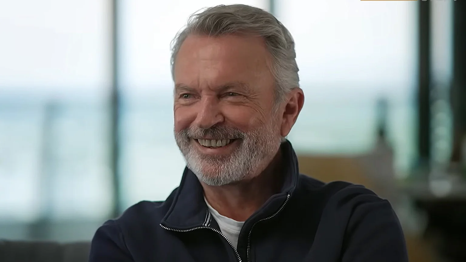 A man with gray hair and a gray beard smiles warmly, wearing a navy blue zip-up jacket, against a softly blurred, light-colored background.
