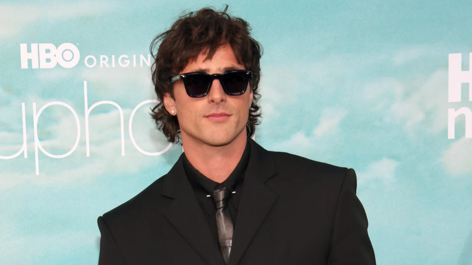 Jacob Elordi, wearing sunglasses, a black suit, and a dark tie, looks forward against a light blue "HBO ORIGINAL Euphoria" background.