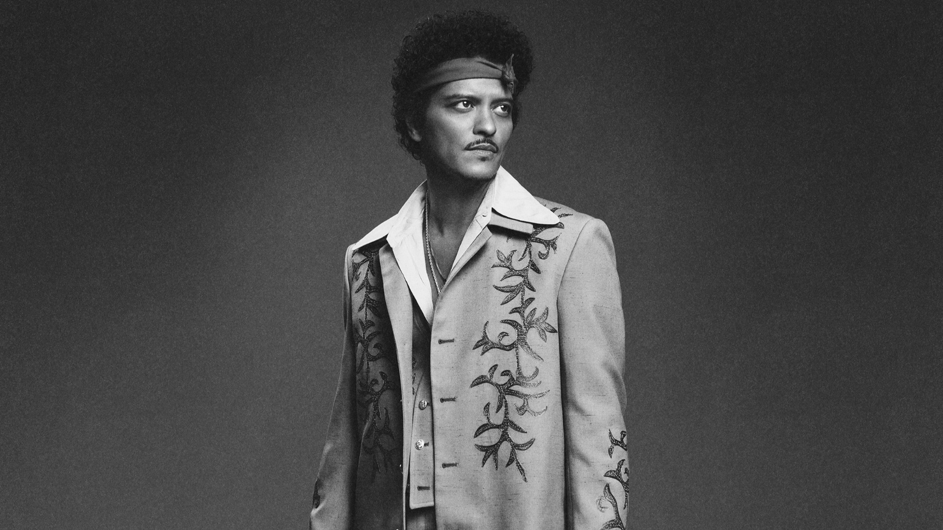 Black and white portrait of Bruno Mars, wearing a patterned jacket over an open white shirt, and a headband, looking to his right.