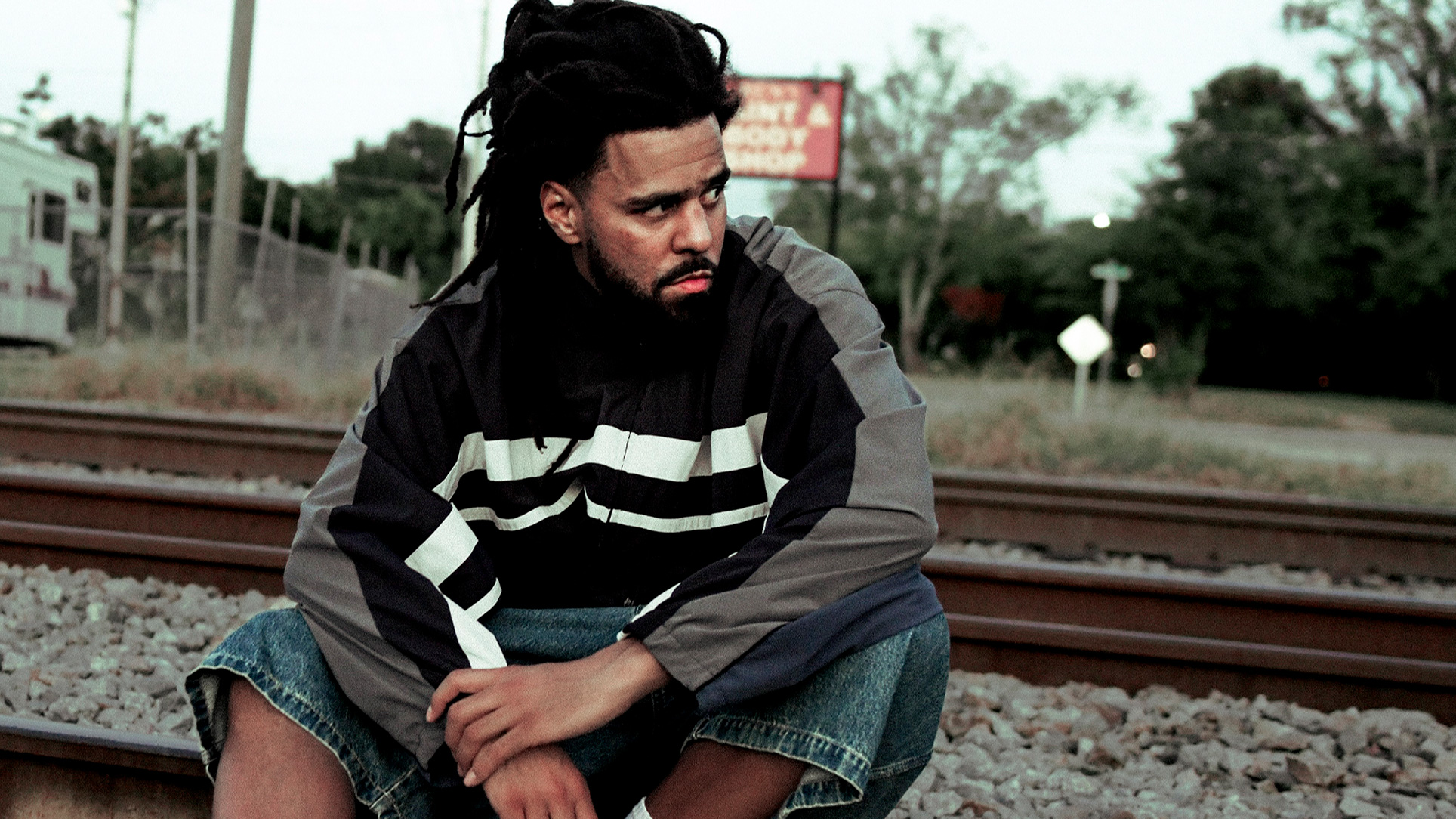 J. Cole, a Black man with dreadlocks and a beard, is crouching on railroad tracks wearing a black, grey, and white windbreaker jacket and blue denim shorts. He looks to the right with a serious expression. The outdoor scene includes a chain-link fence, a white van or trailer, green trees, overgrown grass, and a red sign reading "RENT A BODY SHOP" under an overcast sky.