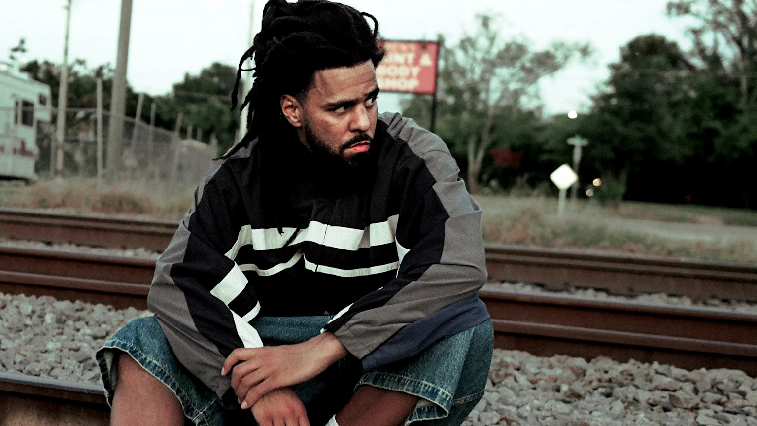 J. Cole, a Black man with dreadlocks and a beard, is crouching on railroad tracks wearing a black, grey, and white windbreaker jacket and blue denim shorts. He looks to the right with a serious expression. The outdoor scene includes a chain-link fence, a white van or trailer, green trees, overgrown grass, and a red sign reading "RENT A BODY SHOP" under an overcast sky.