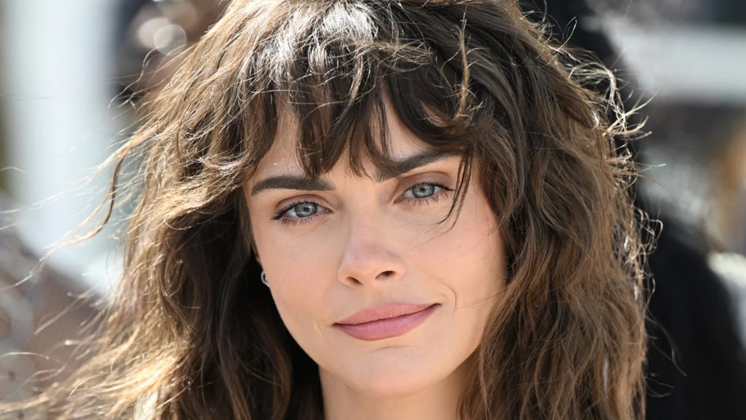 Close-up portrait of a woman with windswept brown wavy hair and bangs, light blue eyes, and a slight smile, looking directly at the viewer.