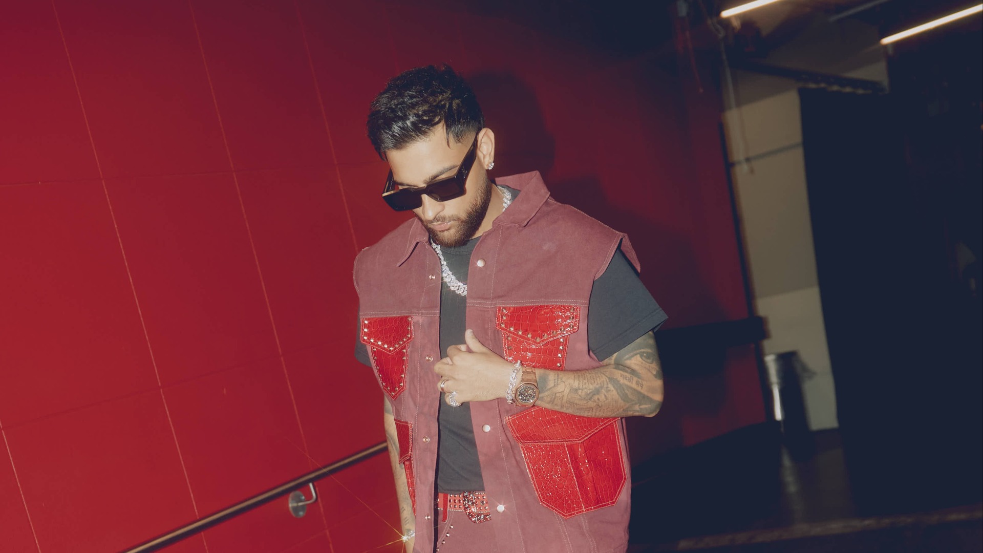 A man with a beard and sunglasses looks down, wearing a reddish-brown studded vest with red pockets over a black t-shirt, a chunky silver chain, watch, and tattoos on his arms, against a red background.