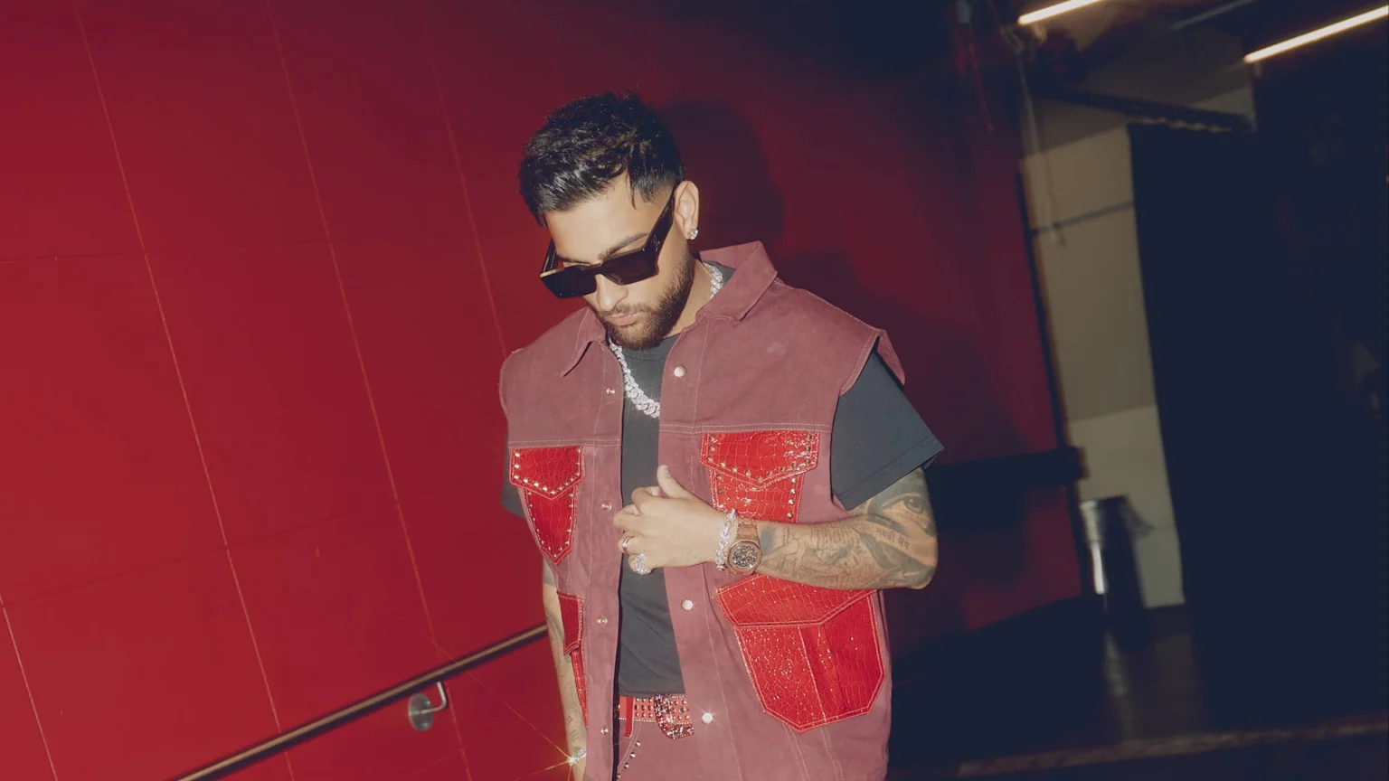 A man with a beard and sunglasses looks down, wearing a reddish-brown studded vest with red pockets over a black t-shirt, a chunky silver chain, watch, and tattoos on his arms, against a red background.