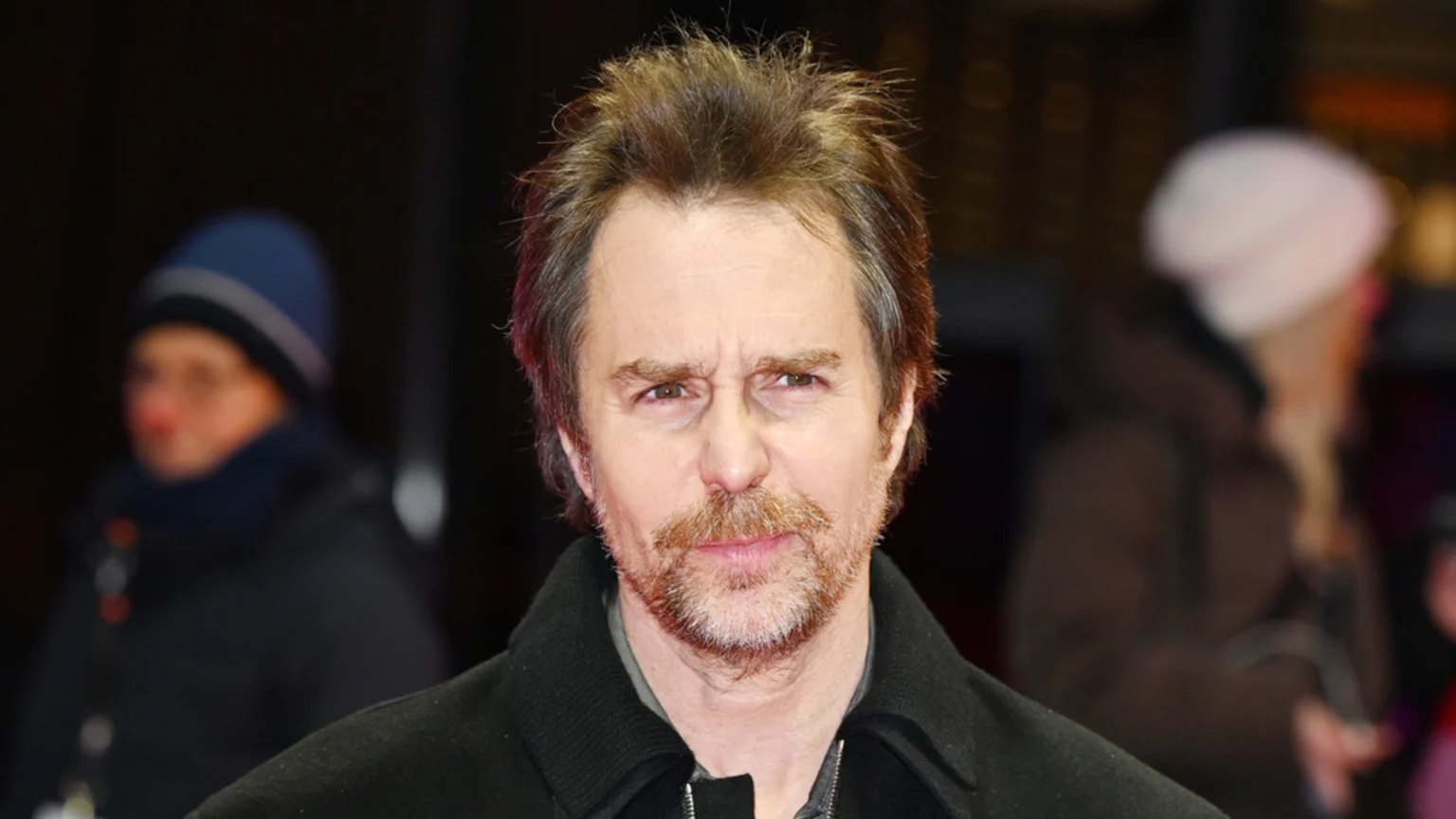A medium close-up portrait of actor Sam Rockwell looking directly at the camera with a neutral expression. He wears a dark jacket and grey shirt against a dimly lit, blurred outdoor public event background with two indistinct figures wearing beanies.