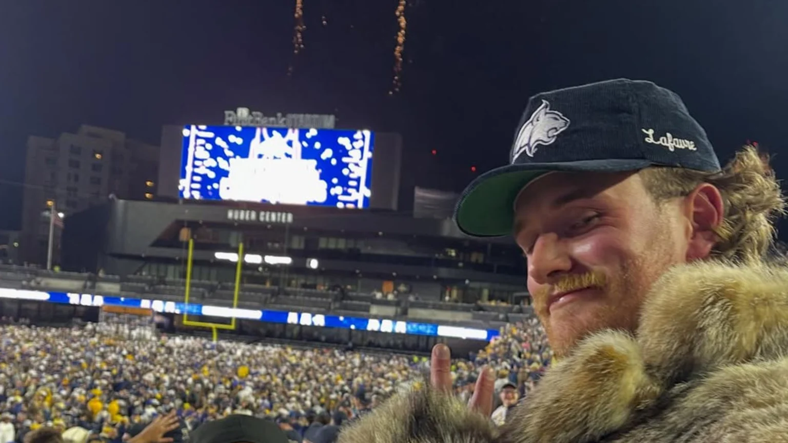 A man with a mustache and mullet, wearing a furry coat and a "Lafaure" cap with a bobcat logo, smiles at a night football game with fireworks and a crowd in the background.
