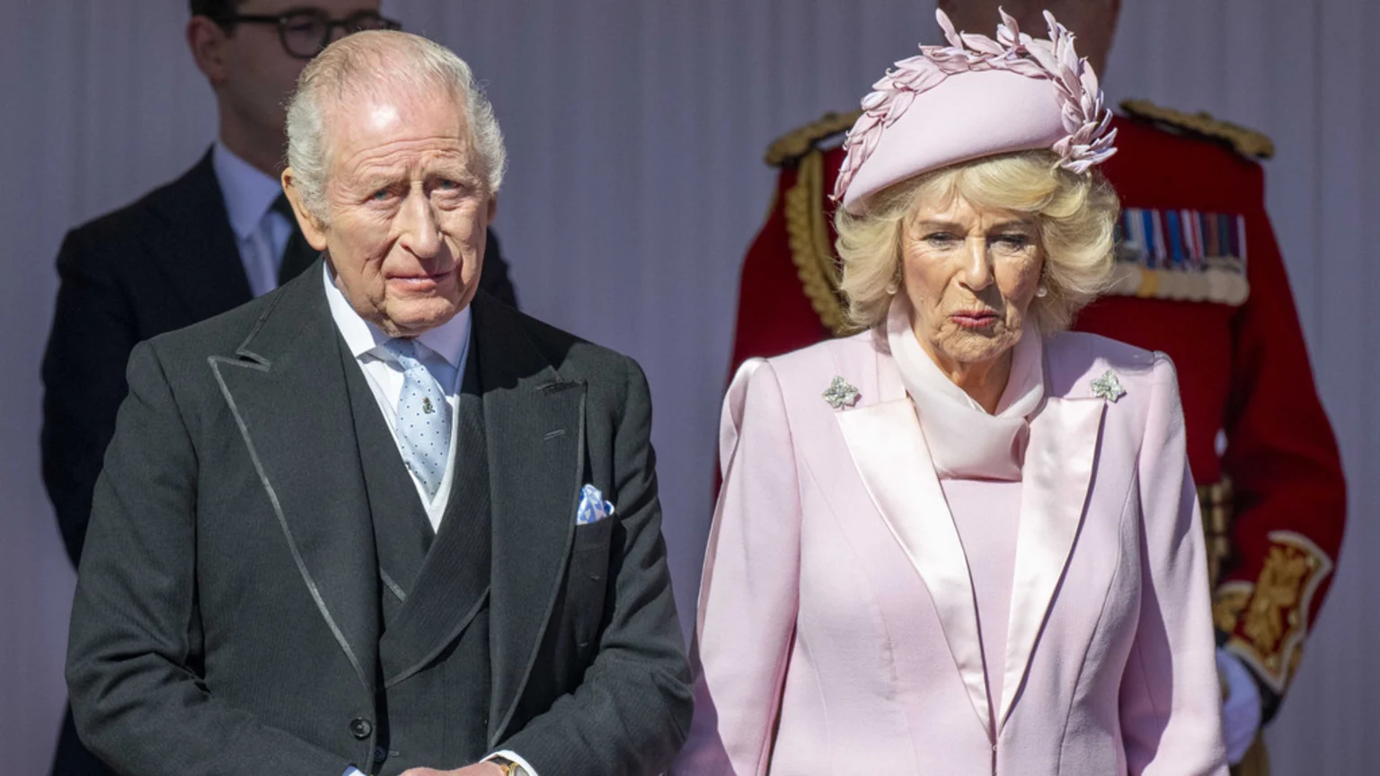 King Charles III in a dark suit and blue tie stands next to Queen Camilla in a pink hat and suit with a brooch, both looking serious.