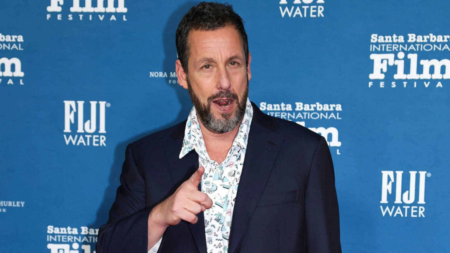 Adam Sandler, with a beard, wearing a dark blazer over a patterned shirt, points forward with his right hand, against a blue backdrop with "Santa Barbara International Film Festival" and "FIJI WATER" logos.