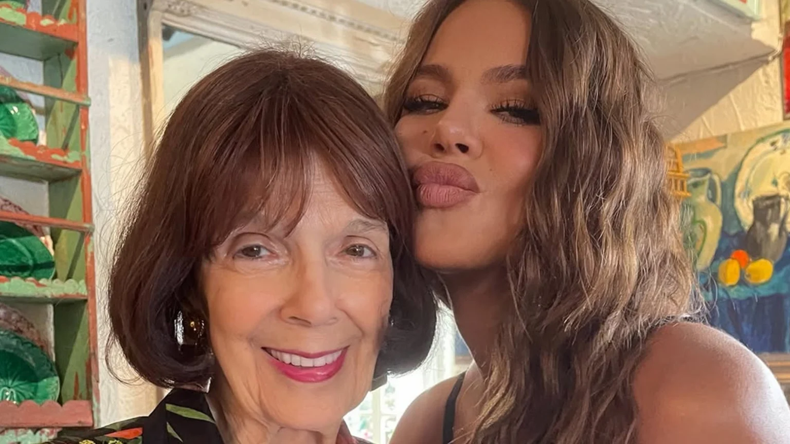 A smiling older woman with brown hair and bangs poses with a younger woman who has long wavy brown hair and is making a kissing face, both looking towards the camera, with a decorative shelf and a still-life painting in the background.