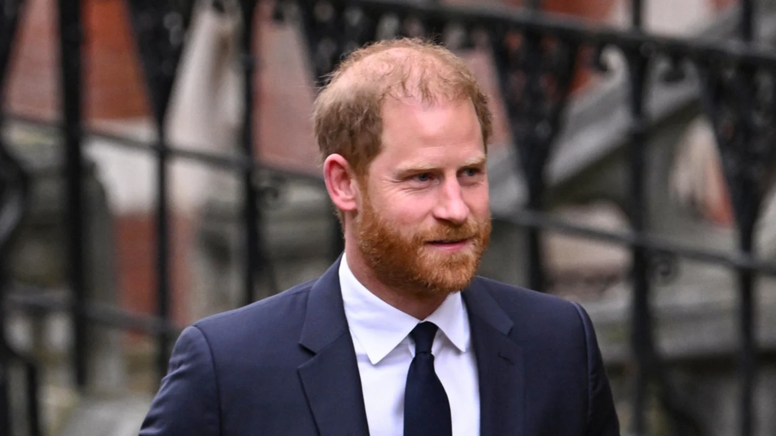 Prince Harry, with reddish-brown hair and beard, smiles slightly while wearing a dark suit, white shirt, and dark tie against a blurred background.