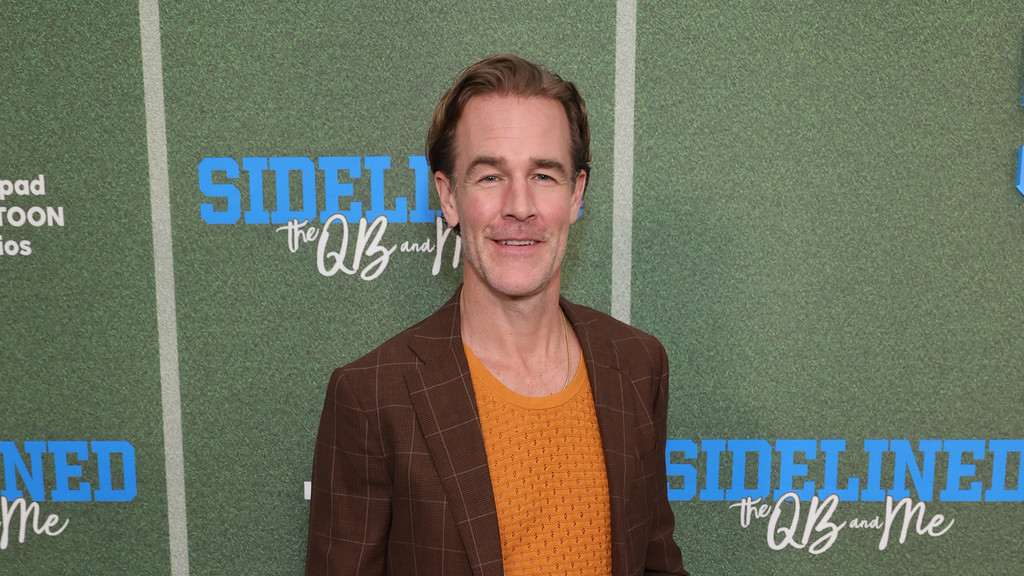 A man with light brown hair and a beard smiles, wearing a brown plaid jacket and an orange shirt, standing in front of a green backdrop with "SIDELINED the QB and Me" text.