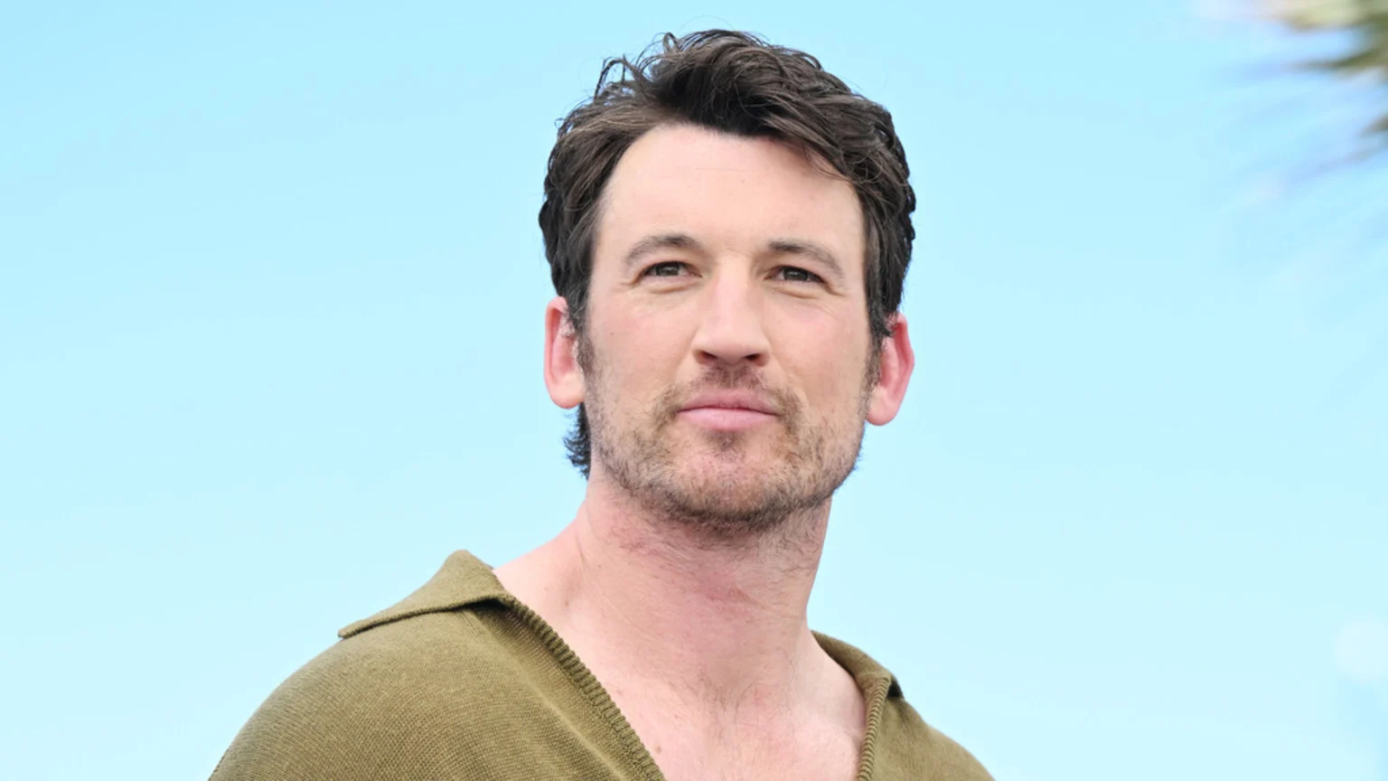 Actor Miles Teller poses against a clear blue sky, wearing an olive-green collared shirt and sporting stubble.