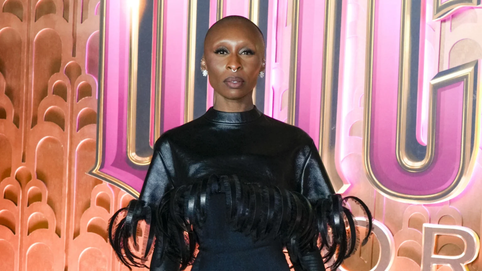 Cynthia Erivo, a woman with a shaved head and nose ring, looks directly at the camera wearing a black top with textured details, in front of pink and gold decorative letters.
