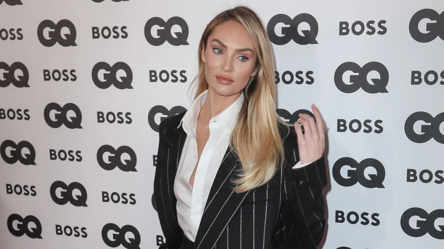 Candice Swanepoel, with long blonde hair, wears a black pinstripe suit and white shirt, her left hand raised, posing against a white backdrop with repeating black 'GQ' and 'BOSS' logos.