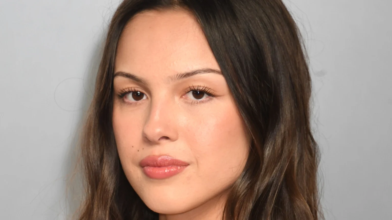 A close-up shot of a young woman with long, dark wavy hair, brown eyes, and natural makeup on a light gray background.