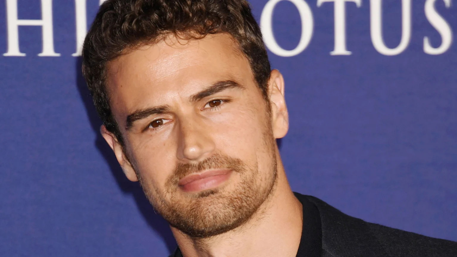 Close-up portrait of Theo James, a man with wavy brown hair, a beard, and brown eyes, looking forward, wearing a dark shirt against a blurred blue background with white text 'THE WHITE LOTUS'.