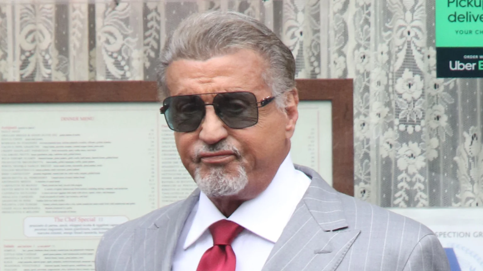 Sylvester Stallone, wearing sunglasses, a grey pinstripe suit, and a red tie, stands in front of a restaurant menu and a lace curtain.