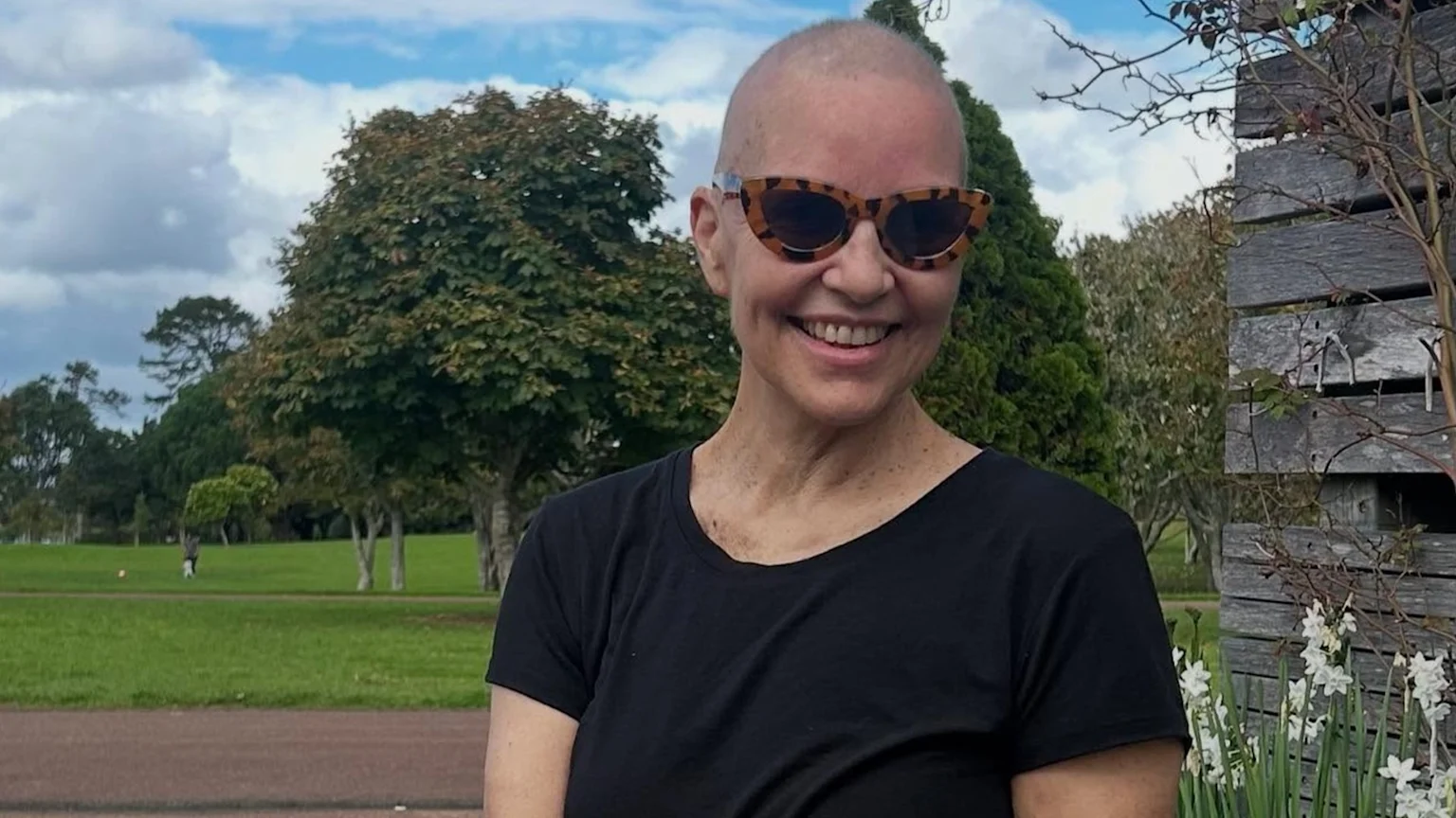 A smiling woman with a shaved head and tortoiseshell sunglasses wears a black t-shirt while standing in a park with trees, a cloudy sky, and a wooden structure.