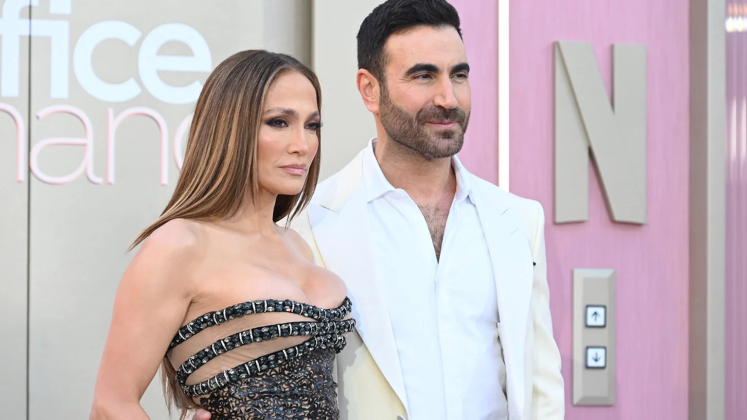 Jennifer Lopez and Brett Goldstein pose together at an event, with Lopez in a strapless dark dress and Goldstein in a white suit, against a backdrop showing "Office Romance" and an "N."