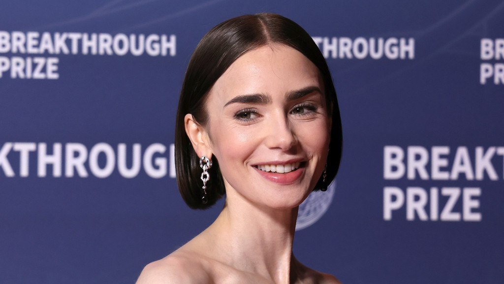 Close-up of Lily Collins smiling, wearing a strapless top and dangling earrings, with a "BREAKTHROUGH PRIZE" banner in the background.
