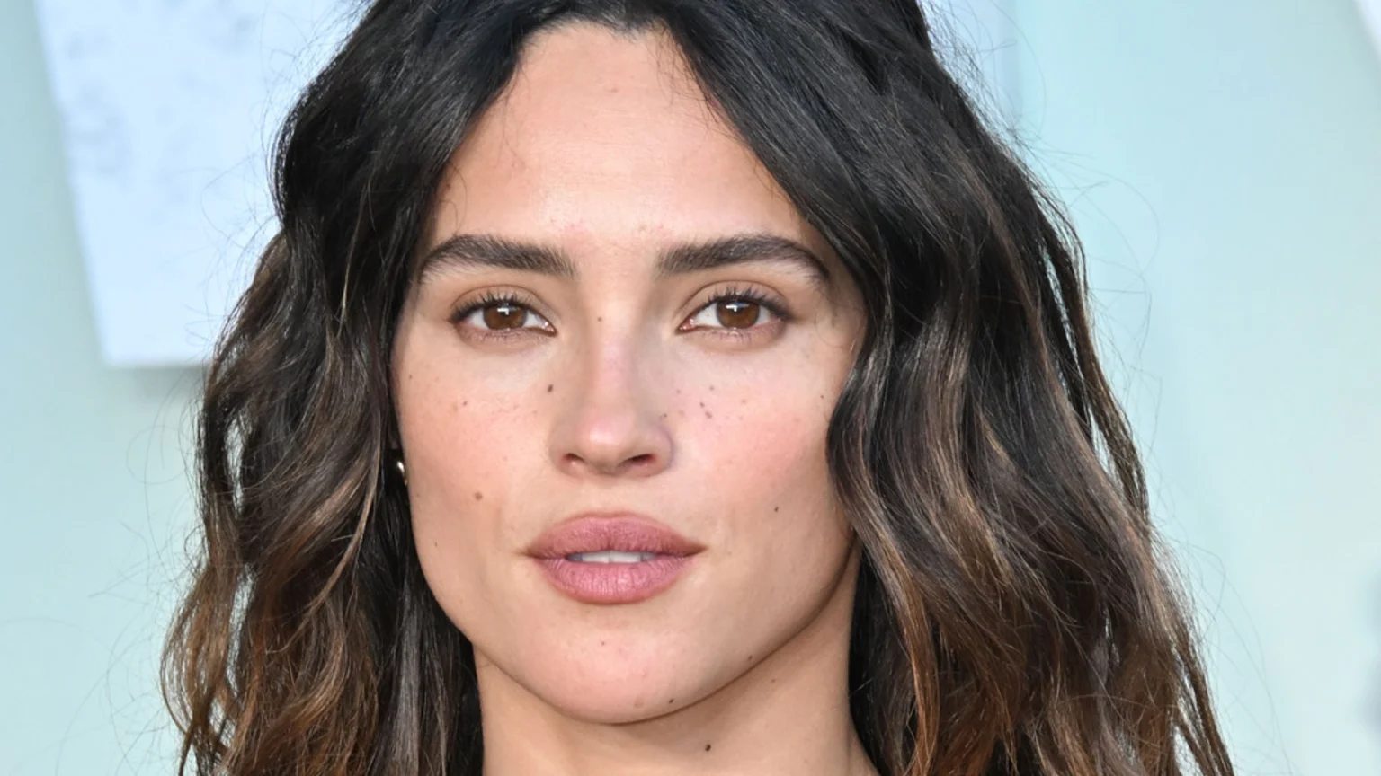 A close-up portrait of a woman with dark, wavy hair, brown eyes, and soft pink lips, looking directly at the camera.