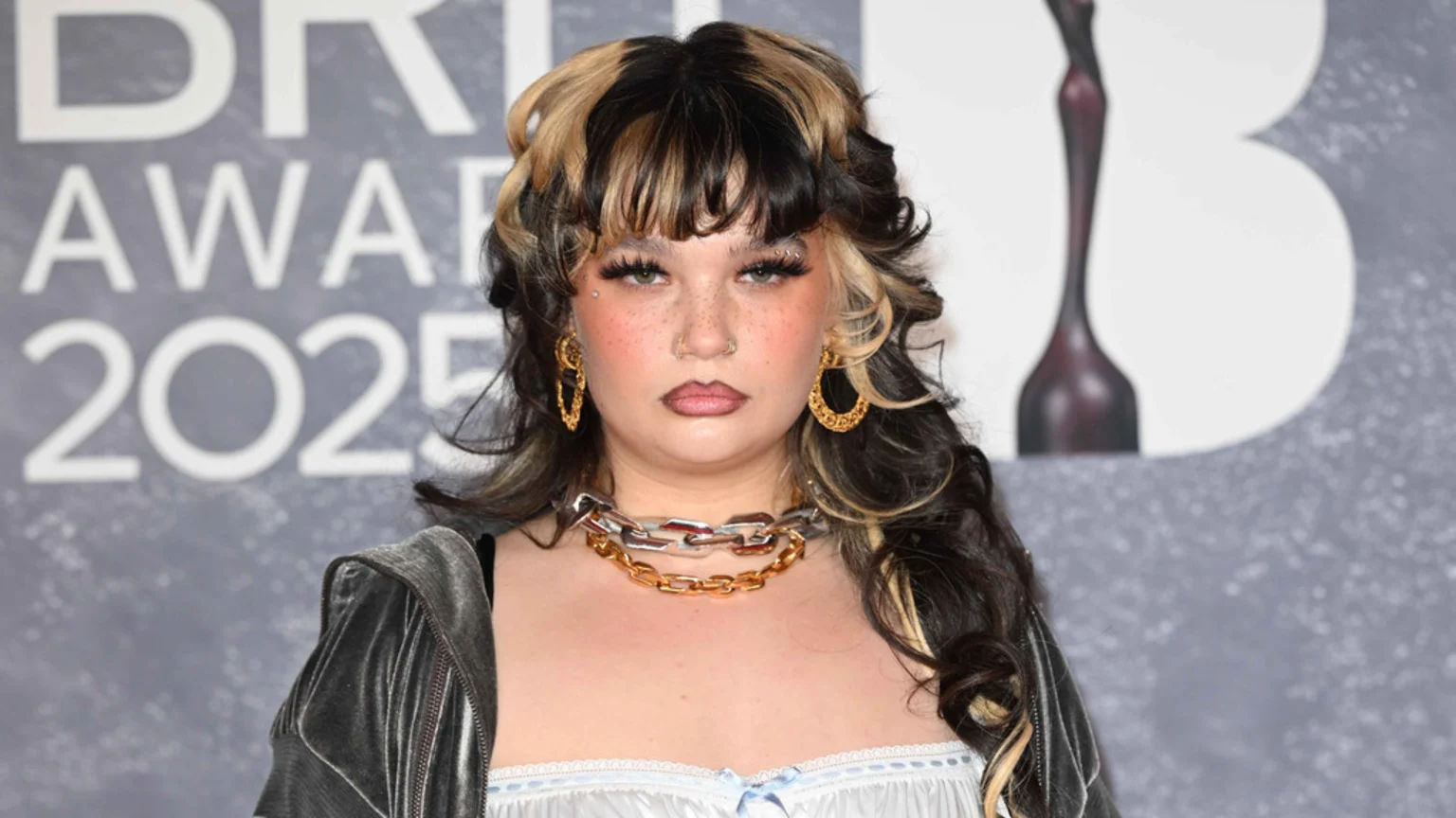 Lola Young, with black and blonde hair and facial piercings, poses at the BRIT Awards 2025 in a dark velvet jacket, layered chain necklaces, and large gold hoop earrings, against a grey background.