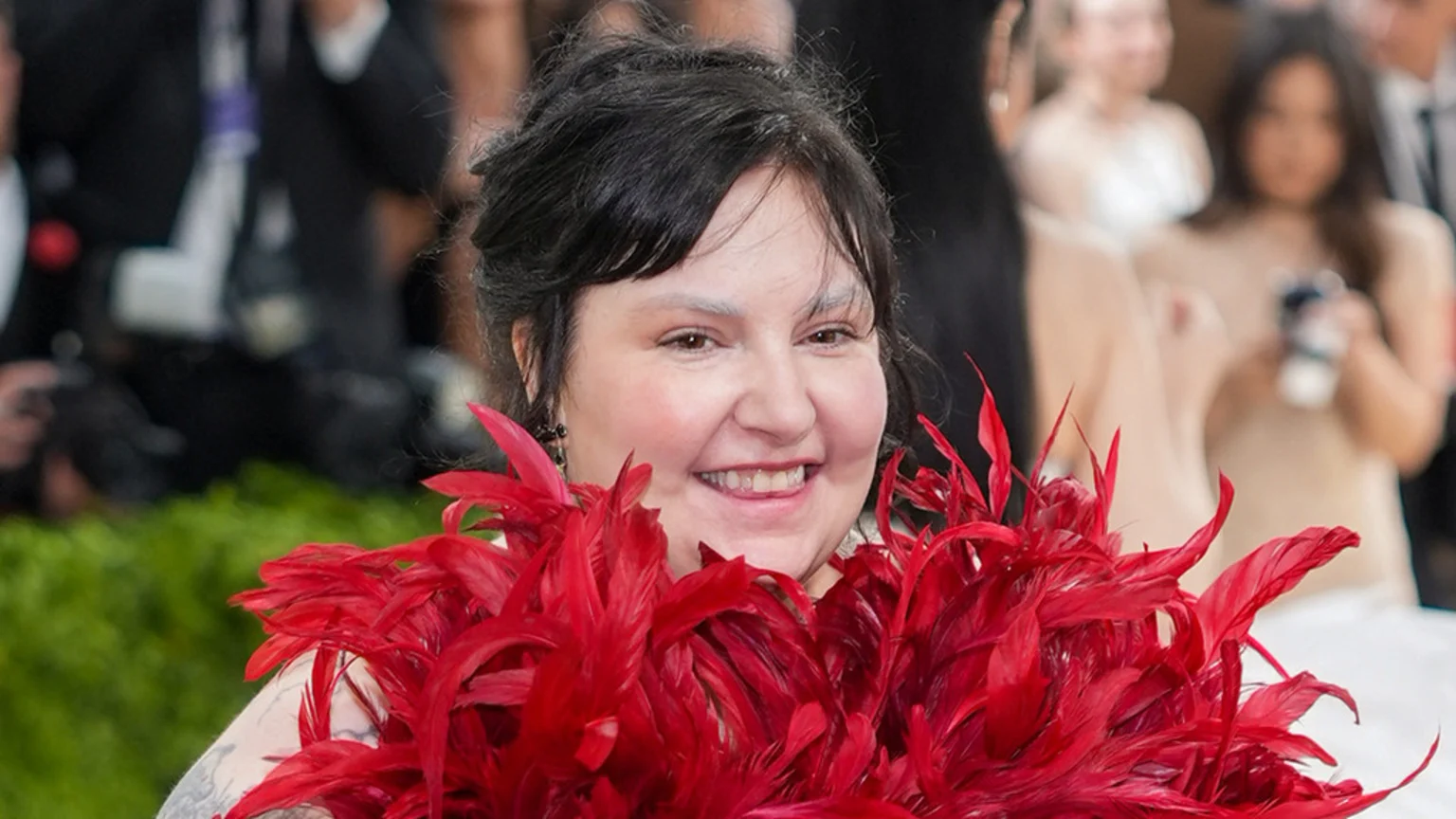 A smiling woman with dark hair, light eyebrows, and visible tattoos wears a vibrant red feathered garment against a blurred background.