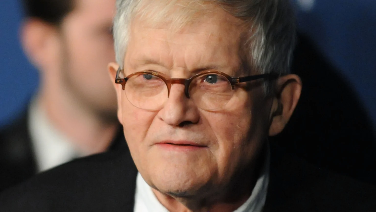 Artist David Hockney, an older man with grey hair and round glasses, wears a dark suit and white shirt, looking forward against a blurred background.
