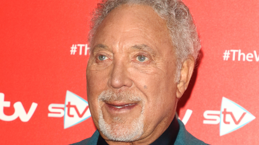 A close-up of an older man with grey, curly hair and a goatee, blue eyes, and a slight smile, standing against a red background with "itv", "stv", and "#TheVoice" logos.