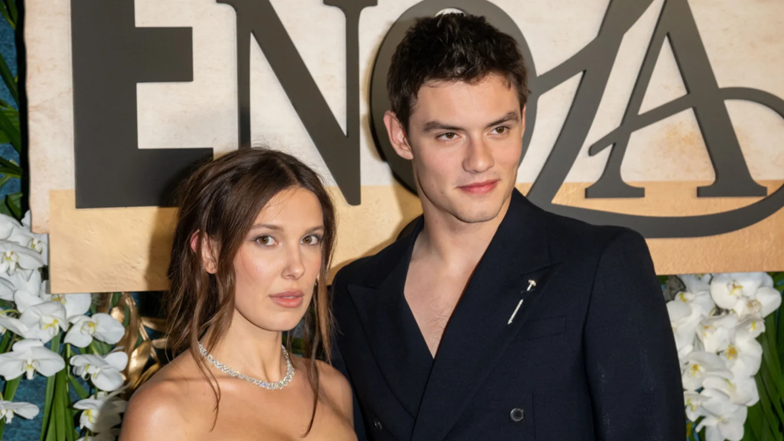 Millie Bobby Brown, wearing a strapless top and diamond necklace, stands next to Louis Partridge, who is in a dark, open suit jacket with a lapel pin, against a backdrop of large letters and white orchids.