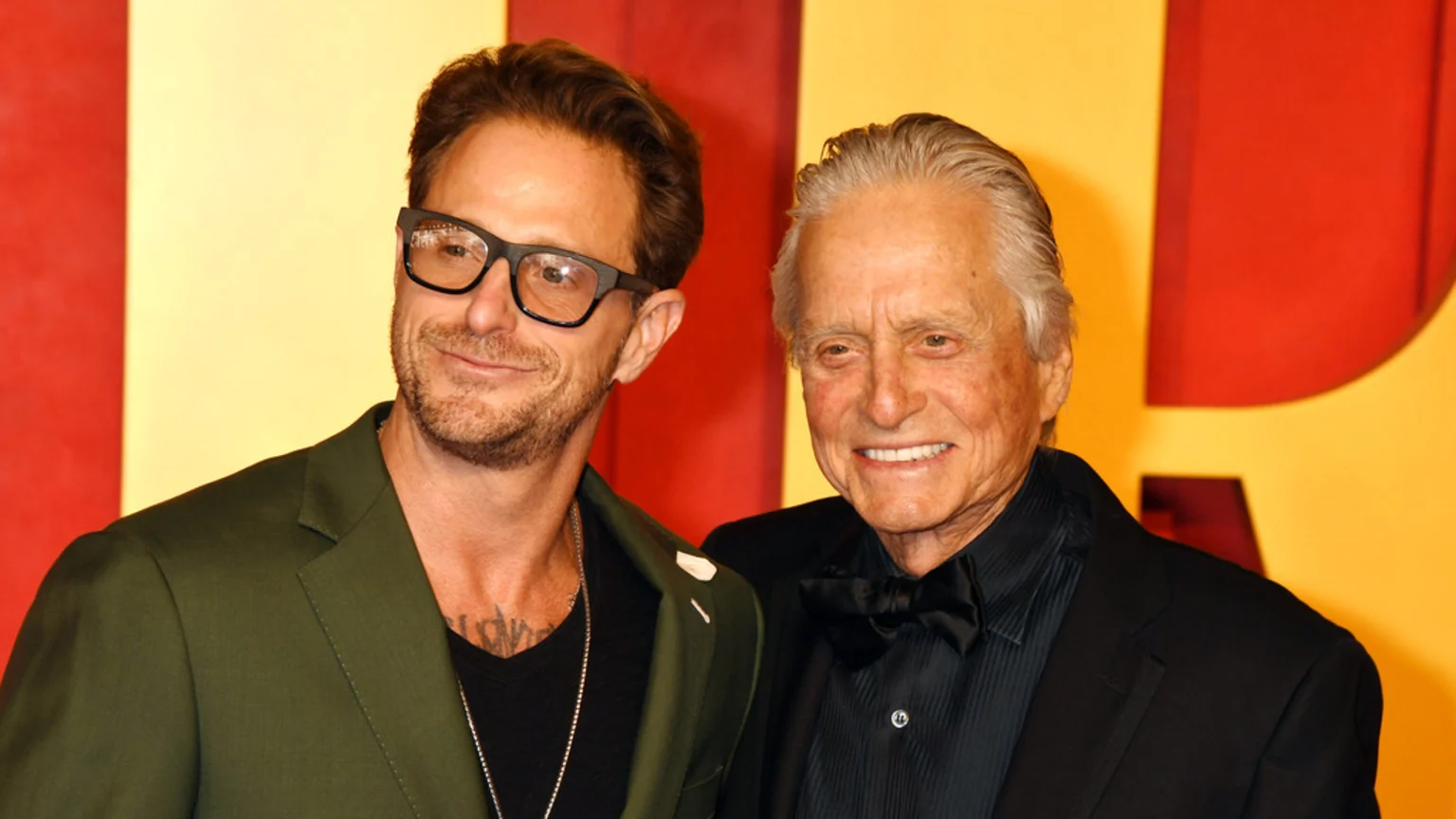 Michael Douglas, in a black suit and bow tie, smiles alongside Rob Reiner, wearing glasses, a beard, and a green blazer, against a yellow and red background.