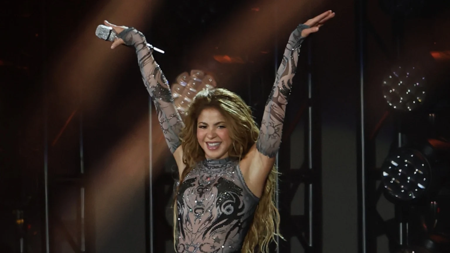 Shakira on stage with arms raised, holding a microphone, smiling broadly while wearing an elaborate, embellished dark top, with stage lights in the background.
