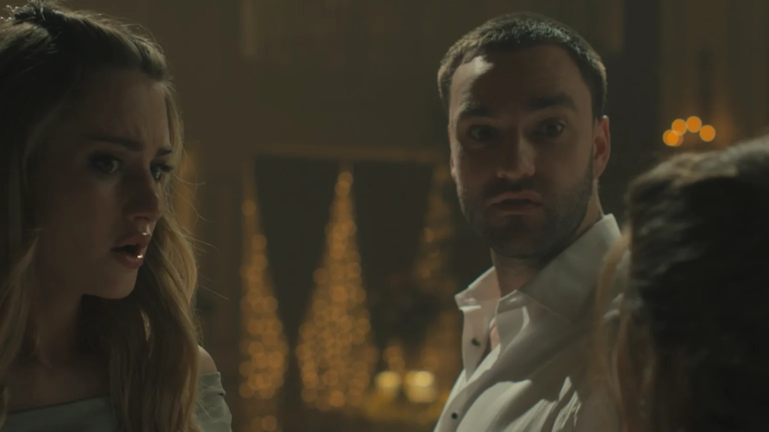 The image shows three unknown individuals at a dimly lit indoor social gathering or event. Two people in the foreground—a woman with blonde hair wearing a light blue/green off-shoulder top and a man with a short beard in a white collared shirt—express surprise or concern. Warm-toned, blurred conical decorative lights resembling Christmas trees illuminate the background, enhancing a festive atmosphere.