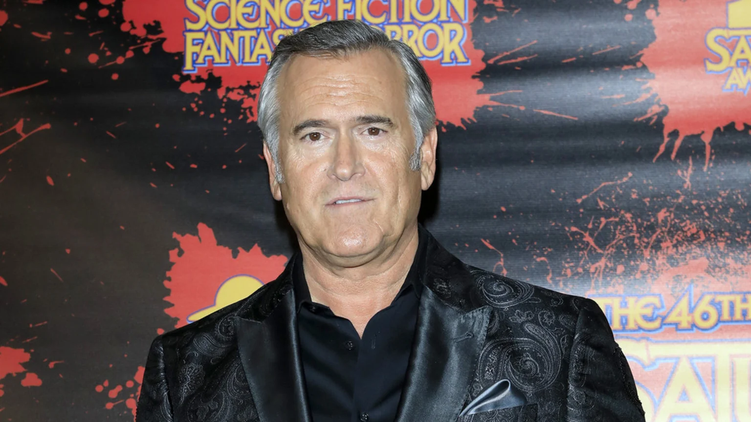 Bruce Campbell, a man with gray hair, wears a black patterned blazer and shirt, looking forward against a dark background with red splatters and event text.