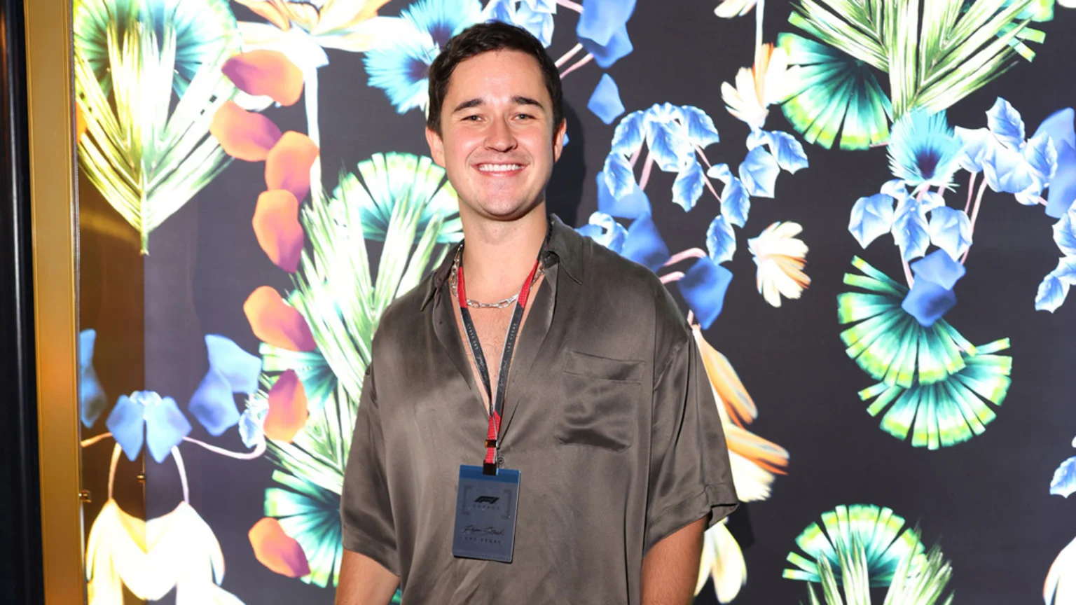 A smiling man identified by his Formula 1 ID badge as John Summit, wearing a dark short-sleeved shirt and silver chain, stands against a vibrant floral background.