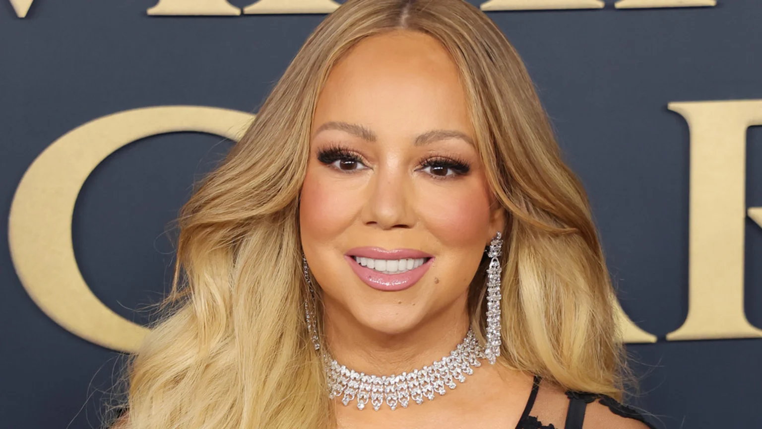 Close-up of Mariah Carey smiling, wearing a diamond choker and earrings, with long blonde hair, against a dark blue background with large letters.